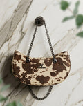 A small crescent-shaped shoulder bag with a brown and beige cow-print pattern.