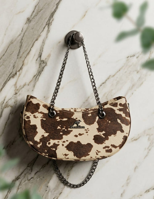 A small crescent-shaped shoulder bag with a brown and beige cow-print pattern.